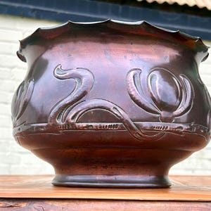 May include: An antique copper planter with a dark patina. The bowl-shaped vessel features ornate, raised floral and swan designs around the exterior. The rim has a scalloped edge. The planter sits on a wooden surface.