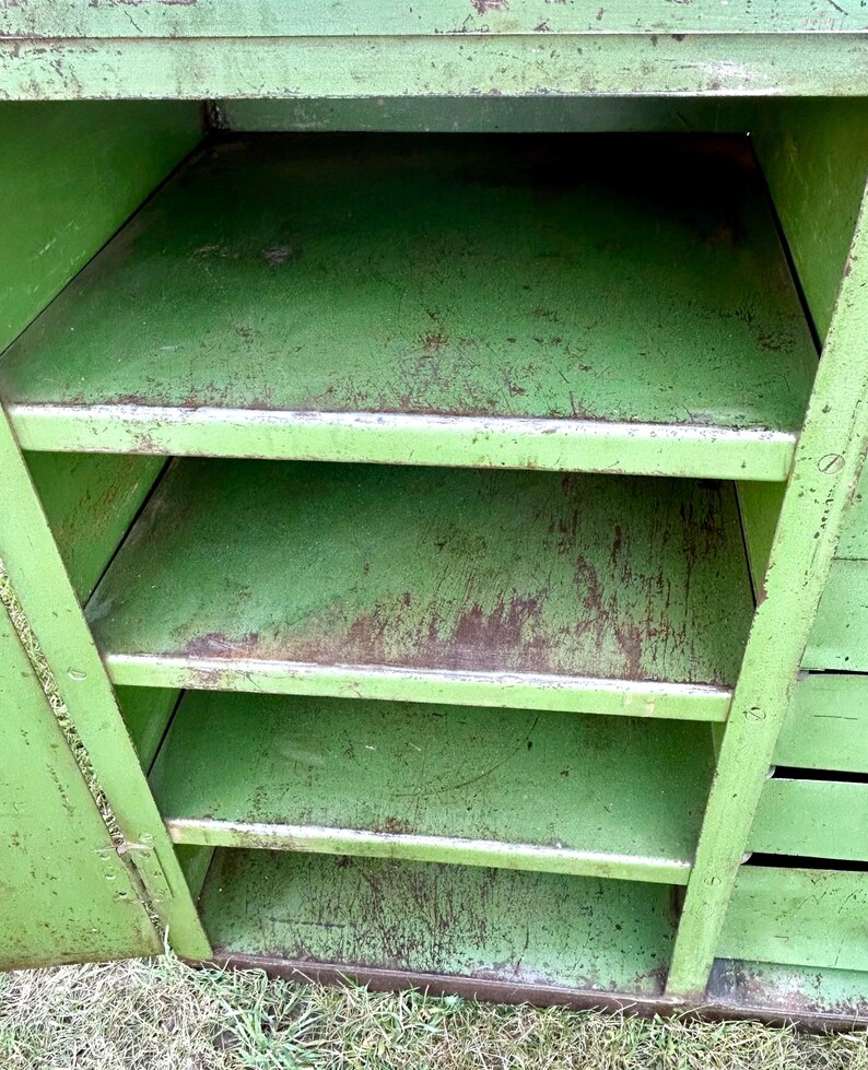 K&ouml;nnte beinhalten: Ein offener, gr&uuml;ner Vintage-Metallschrank mit vier Regalen. Der Schrank hat eine abgenutzte Oberfl&auml;che, die Gebrauchsspuren aufweist. Die Regale sind gleichm&auml;&szlig;ig angeordnet. Der Schrank wird wahrscheinlich zur Aufbewahrung verwendet.