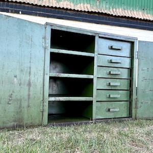 K&ouml;nnte beinhalten: Ein offener, gr&uuml;ner Metallschrank im Vintage-Stil mit f&uuml;nf Schubladen und drei Regalen. Die Schrankt&uuml;ren sind ge&ouml;ffnet und geben den Blick ins Innere frei. Der Schrank steht auf einer grasbewachsenen Fl&auml;che, im Hintergrund ein verwittertes Geb&auml;ude.