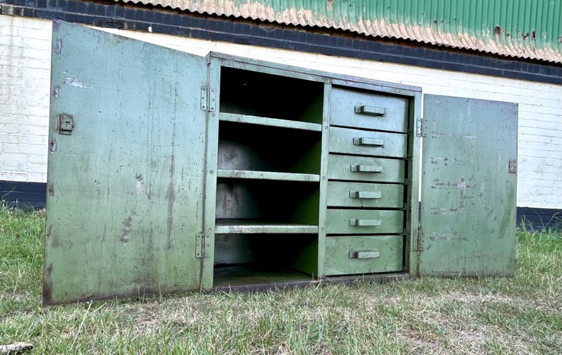 K&ouml;nnte beinhalten: Ein offener, gr&uuml;ner Metallschrank im Vintage-Stil mit f&uuml;nf Schubladen und drei Regalen. Die Schrankt&uuml;ren sind ge&ouml;ffnet und geben den Blick ins Innere frei. Der Schrank steht auf einer grasbewachsenen Fl&auml;che, im Hintergrund ein verwittertes Geb&auml;ude.