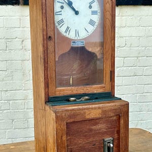May include: Antique wooden time clock with a glass door and a white clock face with black Roman numerals. The clock has a small metal lever on the bottom right side.