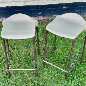 May include: Two off-white metal chairs with a simple design. The chairs have a metal frame and a white plastic seat. The chairs are sitting on a green grassy surface.