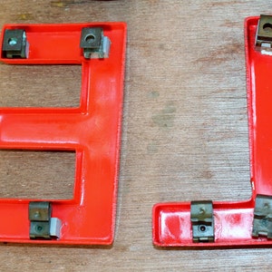 Pu&ograve; includere: Due lettere in plastica rossa, "E" e "L", con elementi di fissaggio in metallo. Le lettere sono su una superficie di legno.