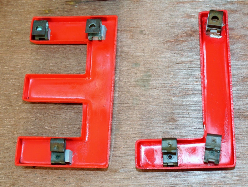Pu&ograve; includere: Due lettere in plastica rossa, "E" e "L", con elementi di fissaggio in metallo. Le lettere sono su una superficie di legno.
