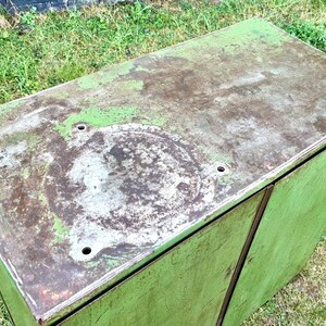 K&ouml;nnte beinhalten: Ein verwitterter, rechteckiger Metallschrank mit verblasstem gr&uuml;nem &Auml;u&szlig;eren. Die obere Oberfl&auml;che weist Abnutzungserscheinungen mit einer gesprenkelten, braun-gr&uuml;nen Patina auf. Es gibt mehrere kreisf&ouml;rmige Vertiefungen und L&ouml;cher auf der Oberseite. Der Schrank steht auf einer grasbewachsenen Fl&auml;che.
