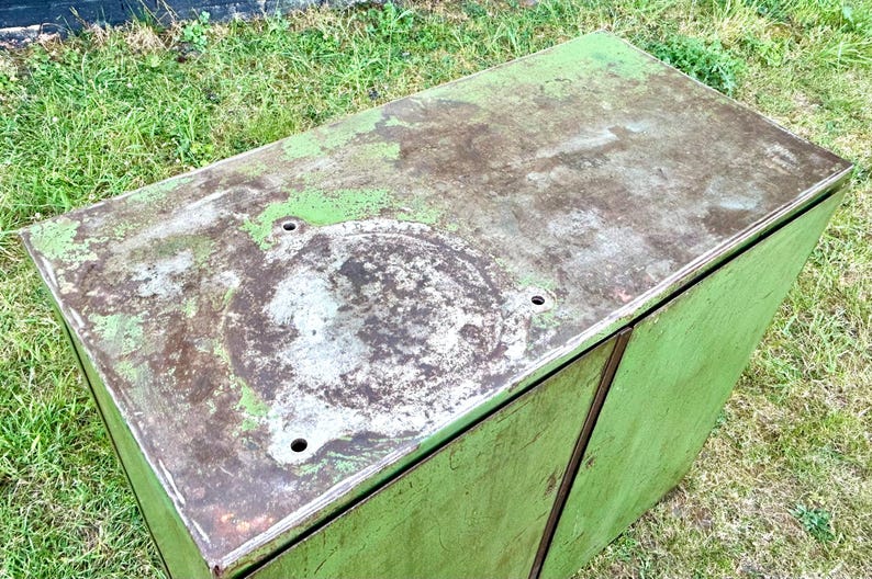 K&ouml;nnte beinhalten: Ein verwitterter, rechteckiger Metallschrank mit verblasstem gr&uuml;nem &Auml;u&szlig;eren. Die obere Oberfl&auml;che weist Abnutzungserscheinungen mit einer gesprenkelten, braun-gr&uuml;nen Patina auf. Es gibt mehrere kreisf&ouml;rmige Vertiefungen und L&ouml;cher auf der Oberseite. Der Schrank steht auf einer grasbewachsenen Fl&auml;che.