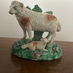 May include: A ceramic sheep and lamb figurine. The sheep stands above the lamb, both with white bodies and brown markings. The base is green with floral details. A decorative ornament, likely for display.