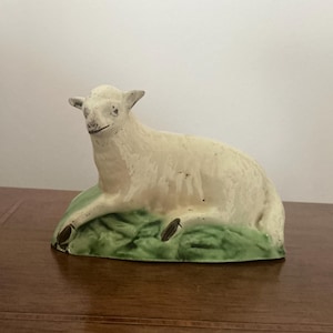 Könnte beinhalten: Eine Keramikfigur eines Lammes, das auf einer grünen Grasfläche liegt. Das Lamm ist cremefarben mit schwarzen Hufen und einem detaillierten Gesicht. Die Figur steht auf einer Holzoberfläche.