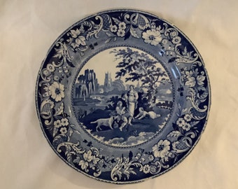 May include: A blue and white porcelain plate with a scenic design featuring a couple and a dog in a landscape with a church and ruins. The plate has a decorative border of floral and scroll motifs.