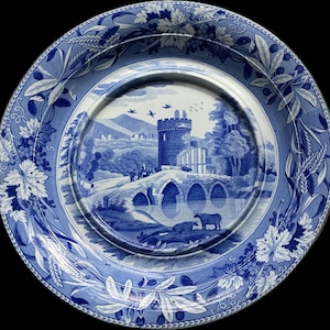 May include: A round ceramic plate featuring a blue and white transferware pattern. The centre depicts a stone bridge, a castle tower, and grazing cattle. The wide rim is decorated with a detailed border of grapevines, wheat, and olive branches.