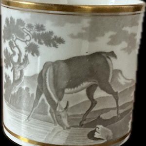 May include: A white ceramic cup with a gold rim and a detailed gray illustration of a deer grazing in a landscape. The design includes trees, hills, and a body of water, creating a pastoral scene. The cup's rim is accented with a thin gold band.