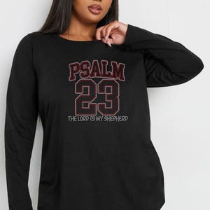 Puede incluir: Camiseta negra de manga larga con el texto "PSALM 23" en un diseño rojo y blanco. El texto "THE LORD IS MY SHEPHERD" está debajo de los números. La camiseta es de material suave.