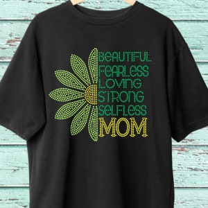 Pode incluir: Camiseta preta com um design de margarida verde e amarela. A margarida tem as palavras "BEAUTIFUL FEARLESS LOVING STRONG SELFLESS MOM" escritas nela.