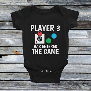 May include: Black baby onesie with white text that reads "Player 3 has entered the game". The text is accompanied by a red, green, and blue joystick graphic.