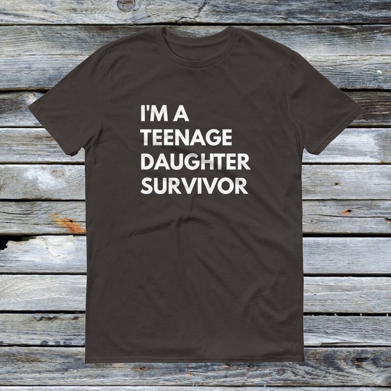 Puede incluir: Una camiseta gris oscuro con el texto "I'm a Teenage Daughter Survivor" impreso en blanco.