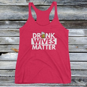 Op de afbeelding: Een rood tanktop met een witte afbeelding waarop "DRUNK WIVES MATTER" staat, met een cartoon-bierglas boven het woord "WIVES".