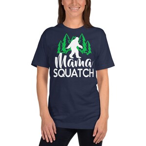 May include: A navy blue t-shirt featuring a white silhouette of Bigfoot walking through a green forest. The text "Mama Squatch" is printed in white below the image.