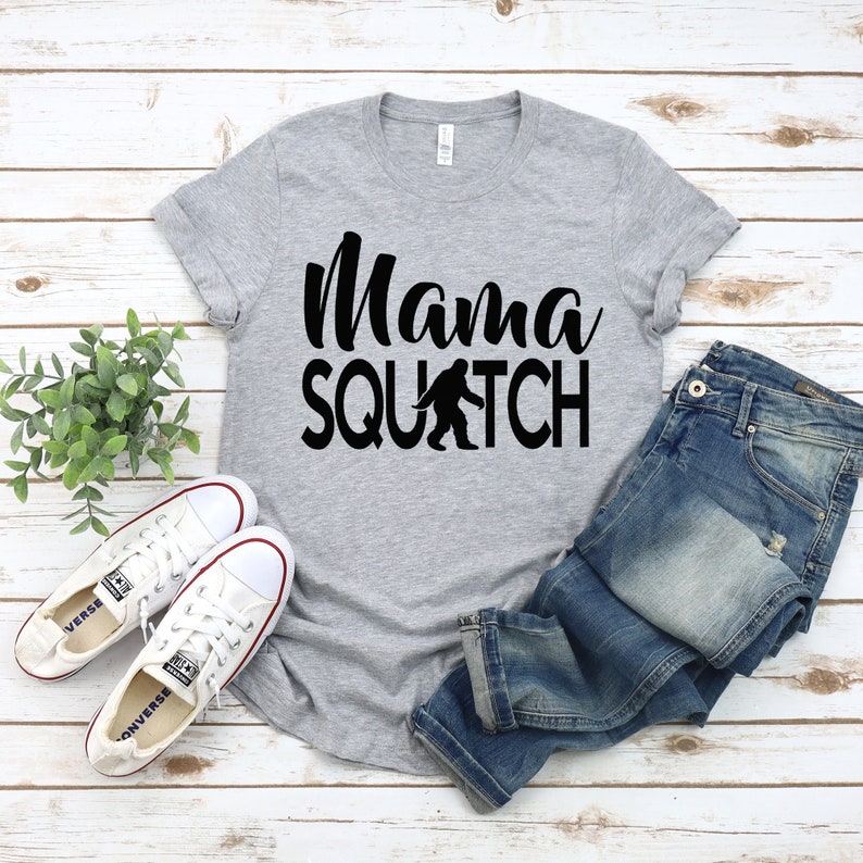 May include: A grey t-shirt with the text "Mama Squatch" in black lettering. A black silhouette of Bigfoot is walking in front of the word "Squatch". The t-shirt is laid out on a white wooden surface with a pair of white trainers and blue jeans.