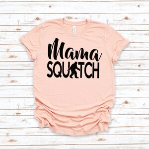 May include: A peach-coloured t-shirt with black text that reads "Mama Squatch" and a black silhouette of Bigfoot walking.