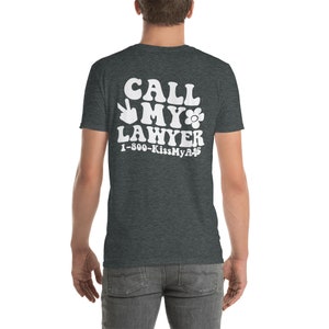 May include: A dark heather grey t-shirt with a white graphic that says "Call My Lawyer" with a peace sign and a flower. The phone number "1-800-KissMyA**" is printed below the graphic.