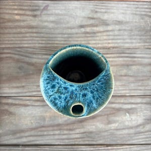 Może przedstawiać: Ręcznie wykonany ceramiczny wazon o unikalnym, asymetrycznym kształcie. Wazon ma żywy niebieski szkliwo o teksturowanym, nakrapianym wyglądzie. Otwór jest owalny, a z boku znajduje się mały otwór. Wazon stoi na drewnianej powierzchni.
