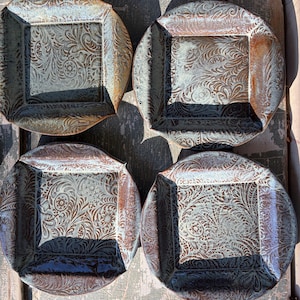 May include: Four square ceramic plates with a decorative floral pattern in shades of brown, blue, and cream. The plates have a slightly raised rim and a glossy finish. The plates are arranged on a weathered wooden surface.