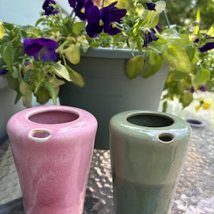 May include: Two ceramic vessels with a smooth, glossy finish. One vessel is a soft pink color with subtle white marbling, while the other is a solid sage green. Each piece features a wide circular opening at the top and a small side hole.