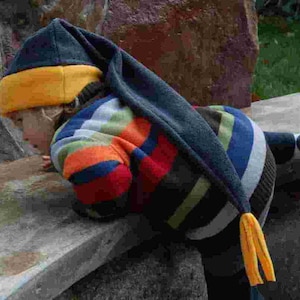 Puede incluir: Un niño con un gorro largo azul oscuro con borde amarillo y un suéter a rayas en tonos azules, naranjas y rojos. El gorro tiene una borla larga. El niño está apoyado en una superficie de piedra.