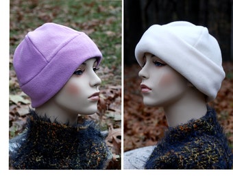 May include: Two fleece hats, one purple and one white, are shown on mannequins. The hats are folded down in the front and have a simple design.