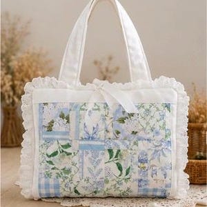 May include: A rectangular quilted tote bag featuring a patchwork design of light blue, green, and white floral and gingham patterns. The bag is bordered by a white ruffled lace trim and includes two top handles and a central white ribbon tie closure.