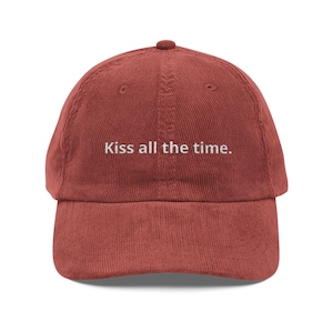 May include: A corduroy baseball cap in a reddish-brown hue. The cap features a curved brim and a rounded top. The phrase "Kiss all the time" is embroidered in white, centered on the front of the cap.