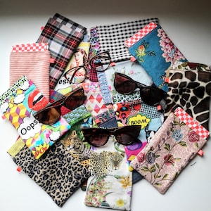 May include: A selection of patterned sunglass cases, featuring floral, leopard print, and geometric designs. Several pairs of sunglasses are visible. The cases are in various colours, including pink, blue, and black. Some cases have a red and white checkered trim.