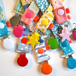 May include: A collection of colorful pacifier clips with various fabric patterns and shapes. Each clip features a different colored button and a metal clip. The fabric patterns include polka dots, geometric shapes, and floral designs.