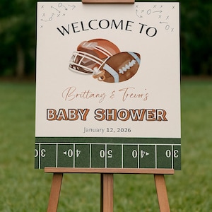 May include: A wooden easel supports a baby shower sign with a football theme. The sign says "WELCOME TO" above a football helmet and ball illustration. Below, it reads "Brittany & Trevor's BABY SHOWER" and "January 12, 2026". The bottom has a green field design.