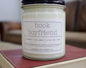 May include: A glass jar candle with a black lid and a white label that reads "book boyfriend." The label also lists the scents: cranberry, cozy amber, silver oak, and musk. The candle is on a red book.