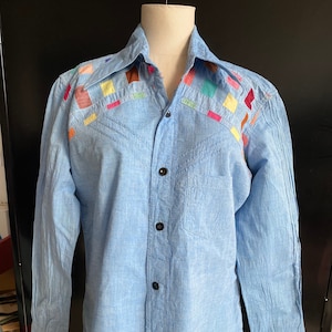 May include: A light blue long-sleeved button-down shirt with colorful patchwork details on the collar and shoulders.