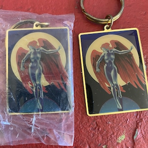 May include: Two gold-colored keychains with a blue background and a winged figure in the center. The figure has red wings and is standing on a blue and white circle. The keychains are on a red surface.