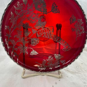 Puede incluir: Un plato redondo de vidrio rojo rubí con un borde plateado festoneado. El plato presenta un diseño floral y el texto "40th Anniversary" en una pancarta. El plato se exhibe sobre un soporte blanco.