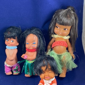 Könnte beinhalten: Vier Vintage-Puppen mit schwarzen Haaren und bemalten Gesichtszügen. Die Puppen tragen bunte Outfits, darunter ein rotes Oberteil mit Tupfen, einen grünen Grasrock und rosa Shorts. Eine Puppe sitzt und scheint zu weinen.