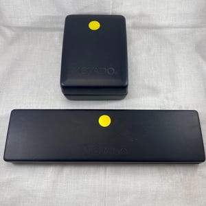 Może przedstawiać: Dwa czarne pudełka na zegarki Movado. Jedno jest prostokątne, a drugie to długi, wąski prostokąt. Oba mają żółtą naklejkę na górze i logo Movado w kolorze białym.