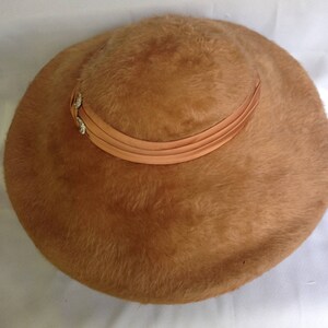 May include: A vintage, round hat in a warm, light brown color. The hat features a soft, fuzzy texture and is adorned with a peach-colored ribbon that encircles the crown. A small, decorative embellishment with rhinestones is attached to the ribbon.