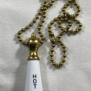 Puede incluir: Una cadena de tracción de latón y porcelana blanca de estilo vintage para una lámpara. El mango de porcelana blanca está inscrito con la palabra "HOT" en letras negras. La cadena está hecha de pequeñas bolas de color latón.