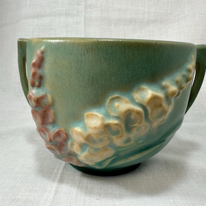 Pode incluir: Uma tigela de cerâmica verde com duas alças e um design floral em tons de creme e rosa. A tigela tem formato arredondado e base verde escura. O design floral é em relevo e adiciona textura.