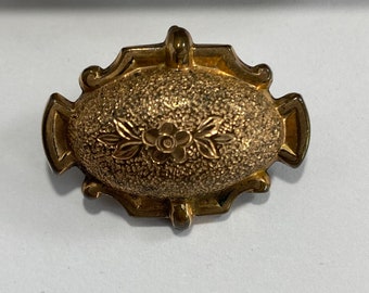 May include: A gold-toned oval brooch with a textured surface and a floral design.