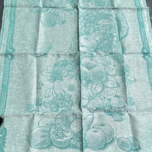 May include: A folded, light green linen towel with a detailed fruit design. The design includes apples, grapes, pears, and pomegranates. The edges are trimmed with a darker green border.