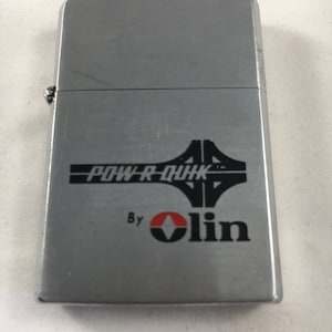 May include: A silver lighter with a black and white graphic of a bridge and the text "POW-R-QUIK" and "By Olin" in black and red.