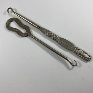 Może przedstawiać: Dwa srebrne metalowe szydełka z ozdobnymi uchwytami. Większe szydełko ma napis "FAIRMONT, MINN." na uchwycie.