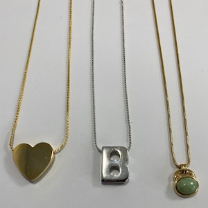 Puede incluir: Tres collares de oro con diferentes colgantes. El primer collar tiene un colgante en forma de corazón. El segundo collar tiene un colgante con la letra B. El tercer collar tiene un colgante redondo de piedra verde.