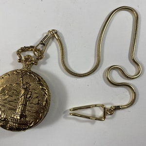 May include: Gold-coloured pocket watch with an embossed Statue of Liberty design. It includes a matching chain and clasp. The watch case is oval with a decorative border. A classic timepiece, ideal for collectors.