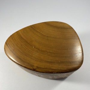 May include: A wooden, triangular-shaped box with a lid. The lid and sides are a warm, light brown color with a wood grain pattern. The box has a smooth, curved design.